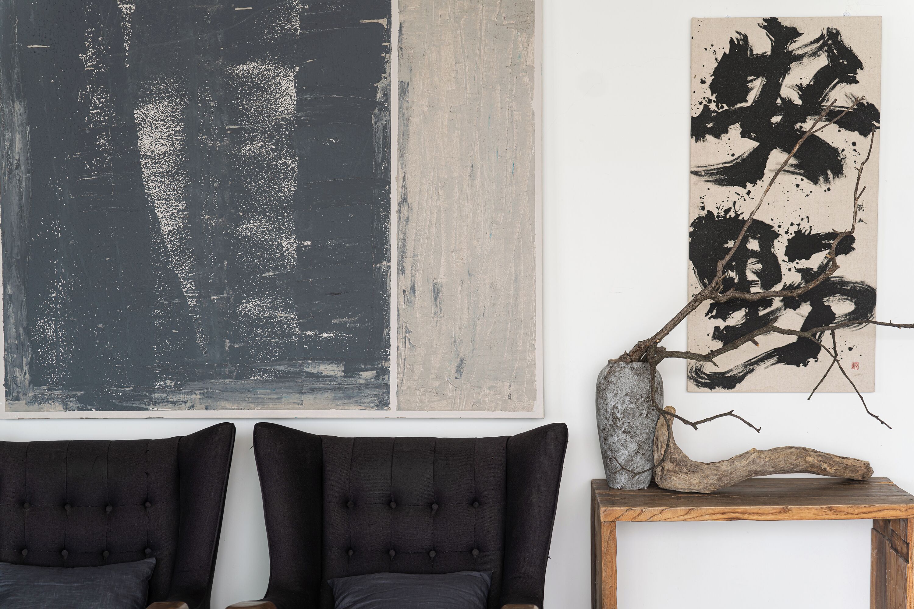 Une pièce comprenant deux fauteuils à l’assise rembourrée de couleur sombre, une table d’appoint en bois et deux grandes peintures abstraites accrochées au mur.
