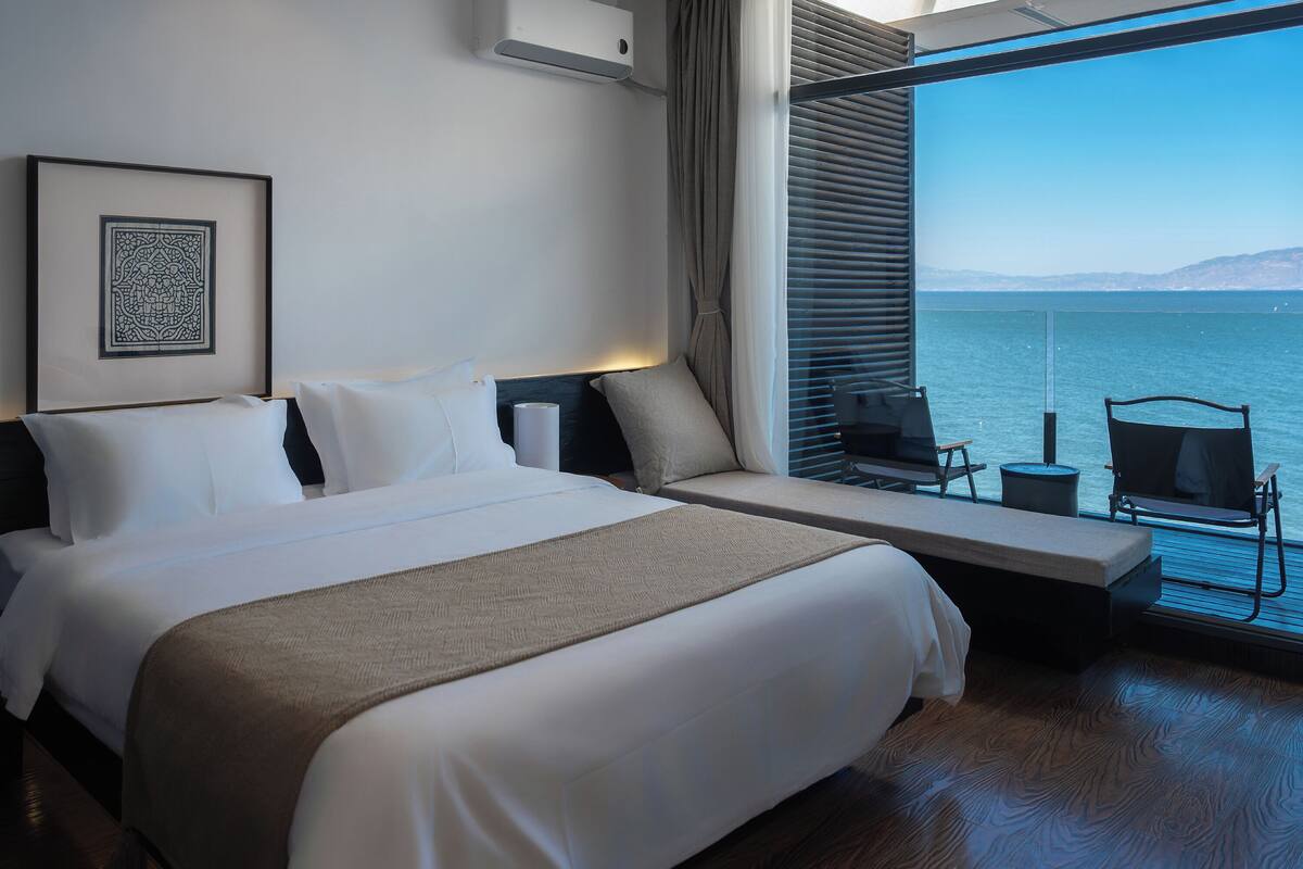 A modern hotel room with a large bed, a bedside table, a chair by the window, and a view of the sea.