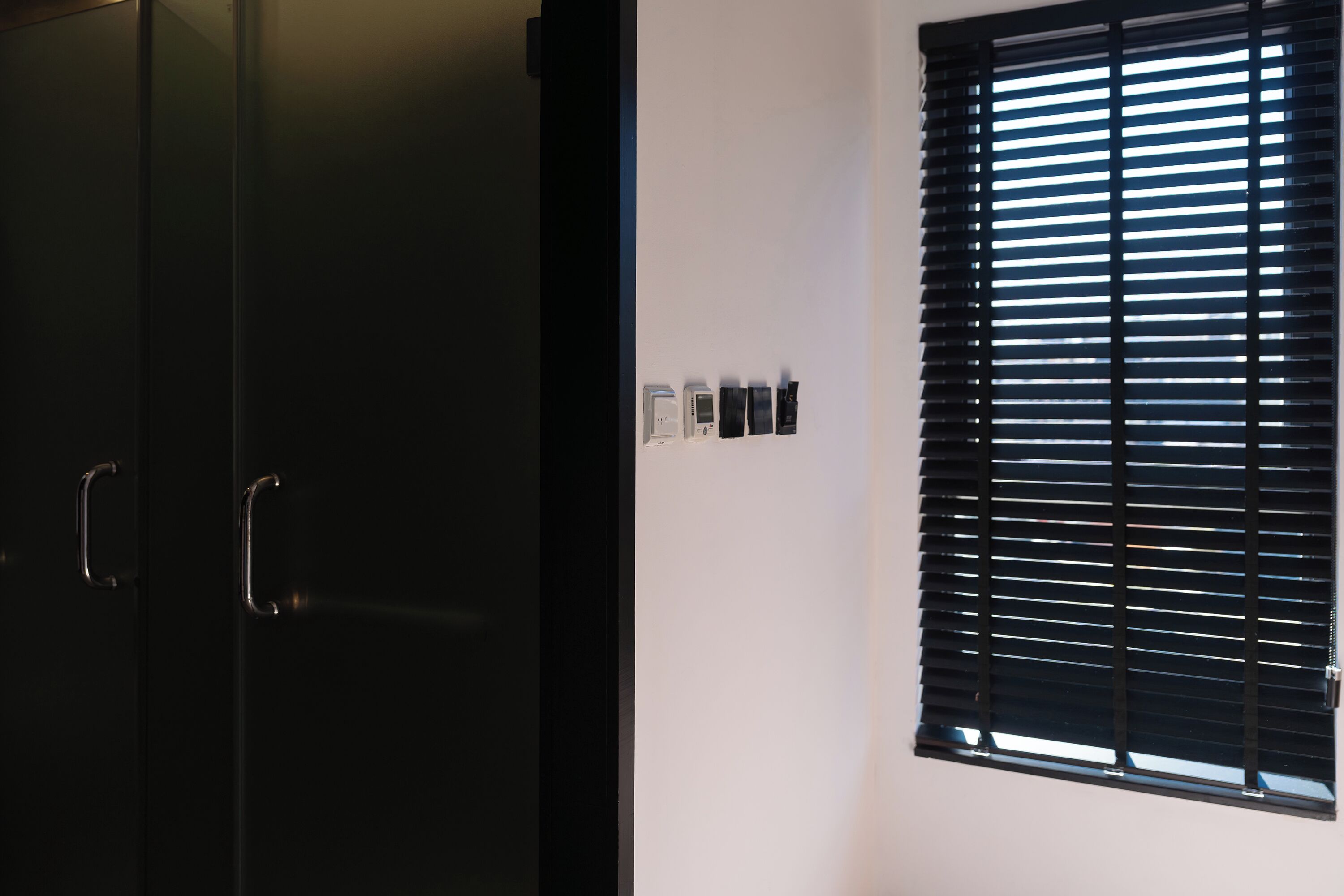 A room with a black wardrobe, a window with horizontal blinds, and a wall-mounted thermostat.