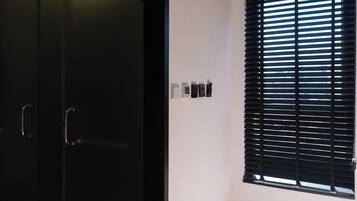 A room with a black wardrobe, a window with horizontal blinds, and a wall-mounted thermostat.