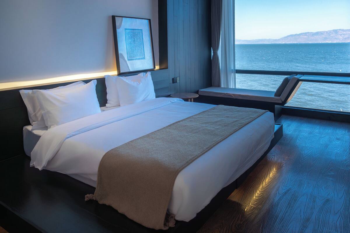 A hotel room with a large bed, a bedside table, a chair, a window with a view of the sea, and a framed picture on the wall.