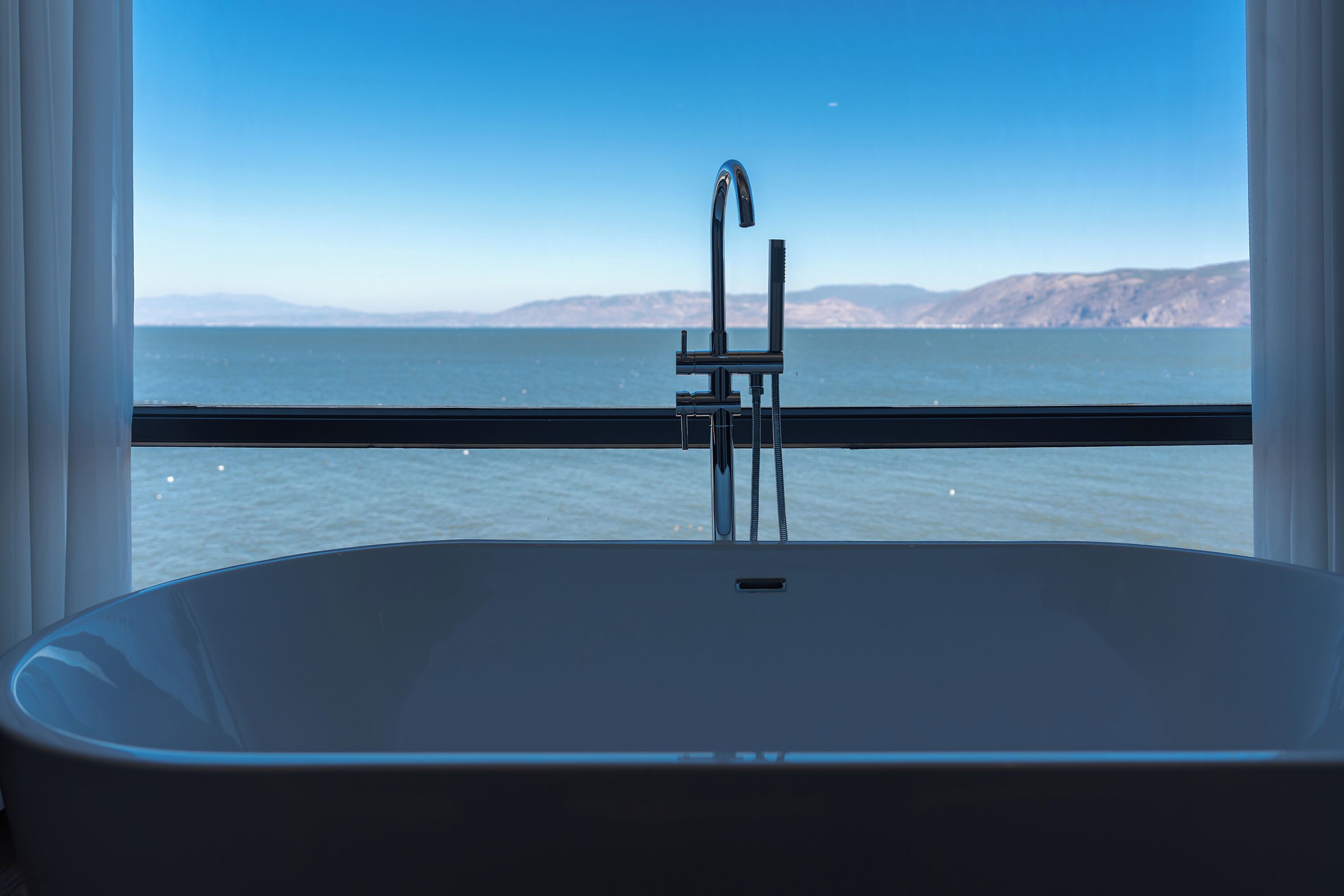 A bathtub with a view of a body of water and distant land through a window.