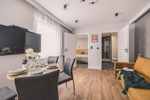 Una habitación moderna con una mesa de comedor lista para cuatro personas, un sofá, un televisor montado en la pared y una puerta que da a un baño.