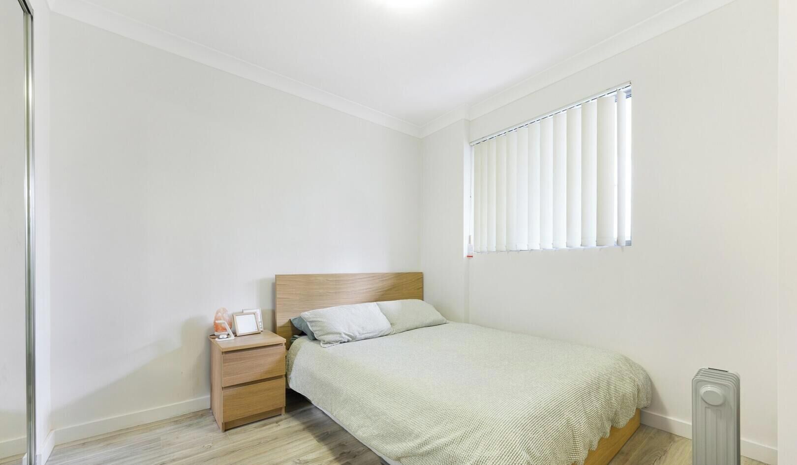 Et lille, rent soveværelse med en enkelt seng, et natbord og et vindue med persienner.