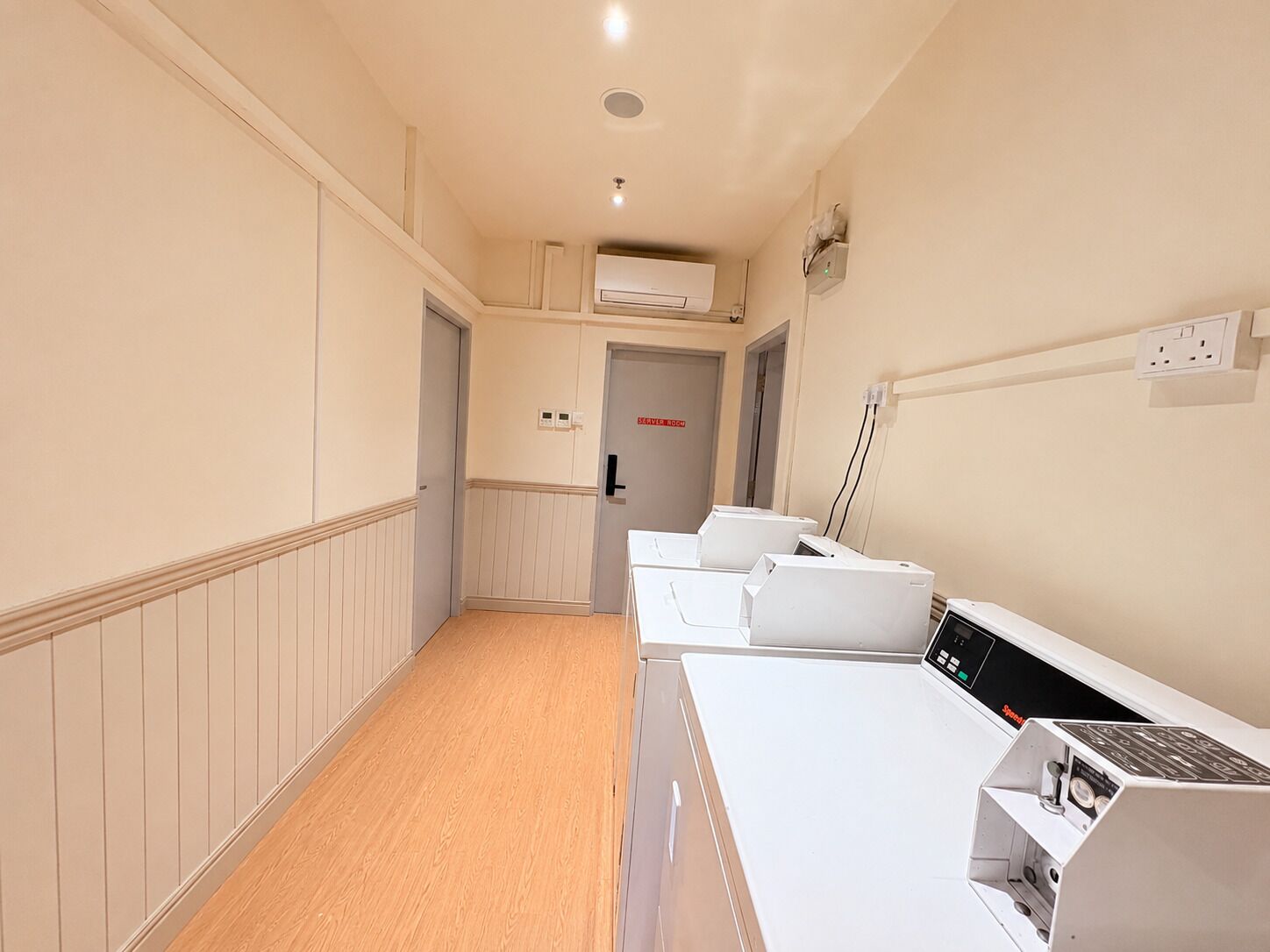 Een gang met een rij witte apparaten, een aan het plafond gemonteerde airconditioner en een deur met de tekst 'SERVICES ROOM'.