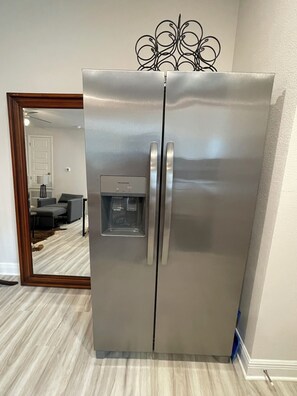 Ein Edelstahl-Kühlschrank mit Wasserspender in einem Raum, der ein Holzrahmen-Spiegel, ein graues Sofa und einen Teppich mit Muster aufweist.