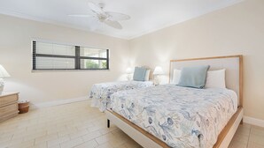 Ein Schlafzimmer mit zwei Betten, einem Deckenventilator, einem Fenster mit Jalousien und einem Nachttisch mit Lampe.