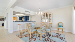 A dining area with a round table, wicker chairs, a patterned rug, and a kitchen in the background. - Casa Caribe #16 - 3 Bed, 3 Bath - Panorama Ocean Front Condo on the Second Floor (Seven Mile Beach)
