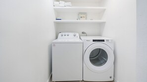 A compact laundry area with a washer and dryer, a shelf with folded towels, and a children's book. - Casa Caribe #24 - 3 Bed, 3 Bath - Third Floor Ocean Front Panorama Condo (Seven Mile Beach)