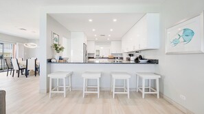 A modern kitchen with white cabinetry, a central island with stools, and a dining area with chairs. - Casa Caribe #24 - 3 Bed, 3 Bath - Third Floor Ocean Front Panorama Condo (Seven Mile Beach)