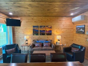 Interior acogedor de una cabaña con paredes de madera, un televisor de pantalla plana, dos cuadros, un sofá y sillones de cuero.