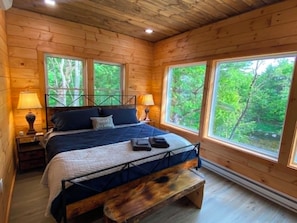 Ein Schlafzimmer einer Holzhütte mit einem Bett, zwei Nachttischlampen, einer Bank und großen Fenstern.