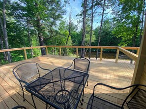 Una terraza de madera con una mesa y sillas de metal, rodeada de árboles y un río.