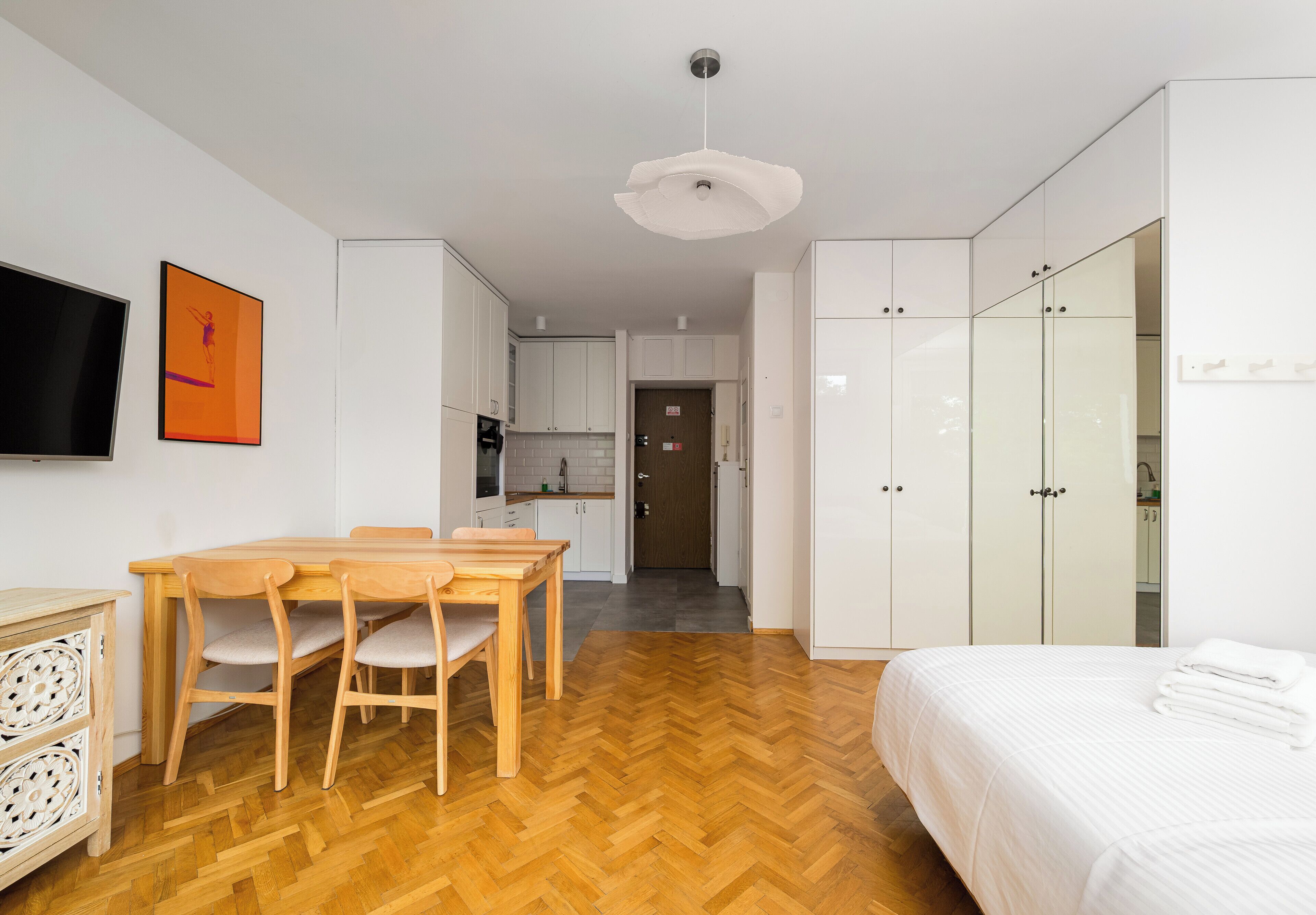 Une chambre moderne avec une table à manger en bois, un lit avec une literie blanche et un coin cuisine à l’arrière-plan.
