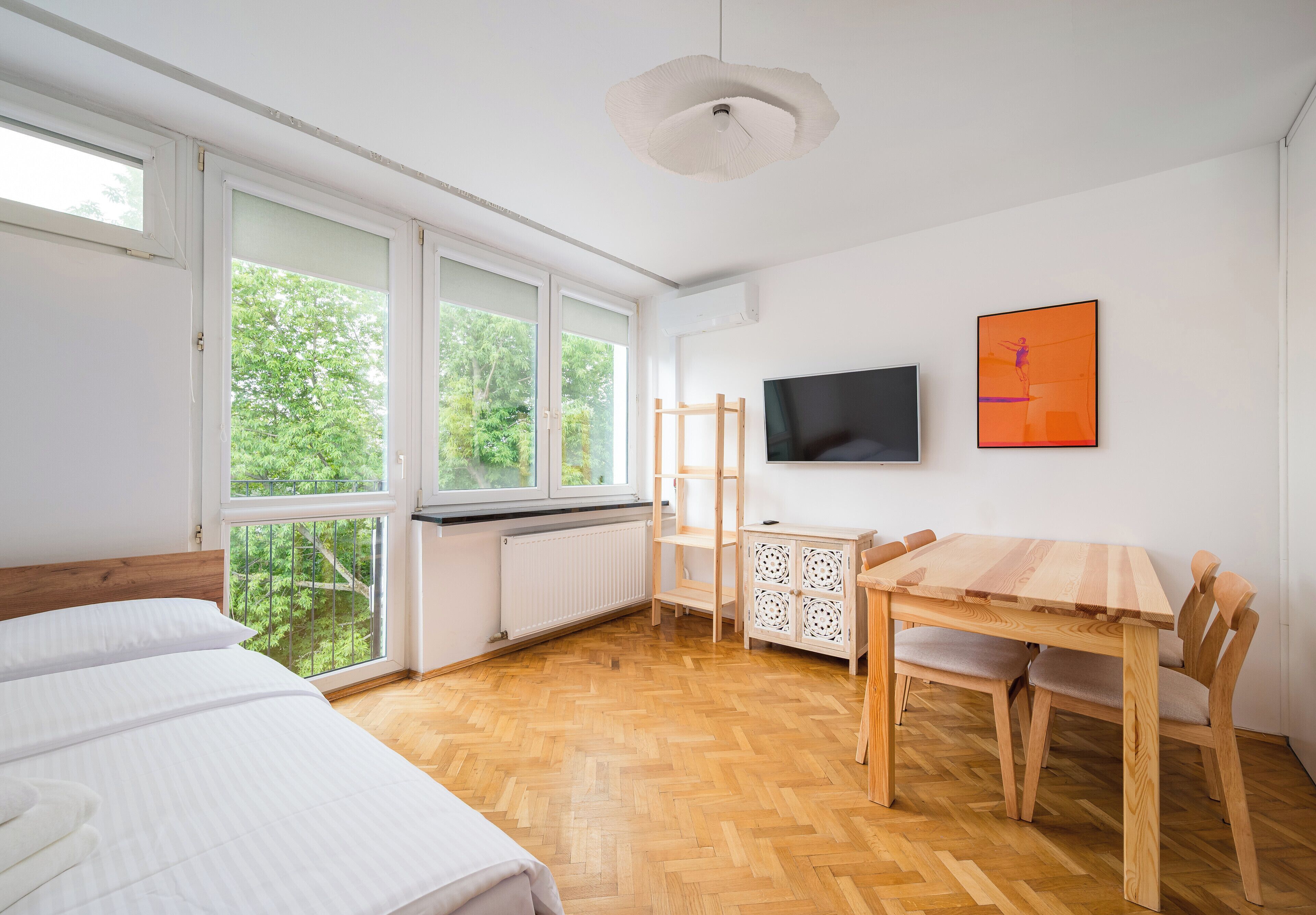 A room with a bed, a wooden dining table with chairs, a television on the wall, and a window with a view of greenery.