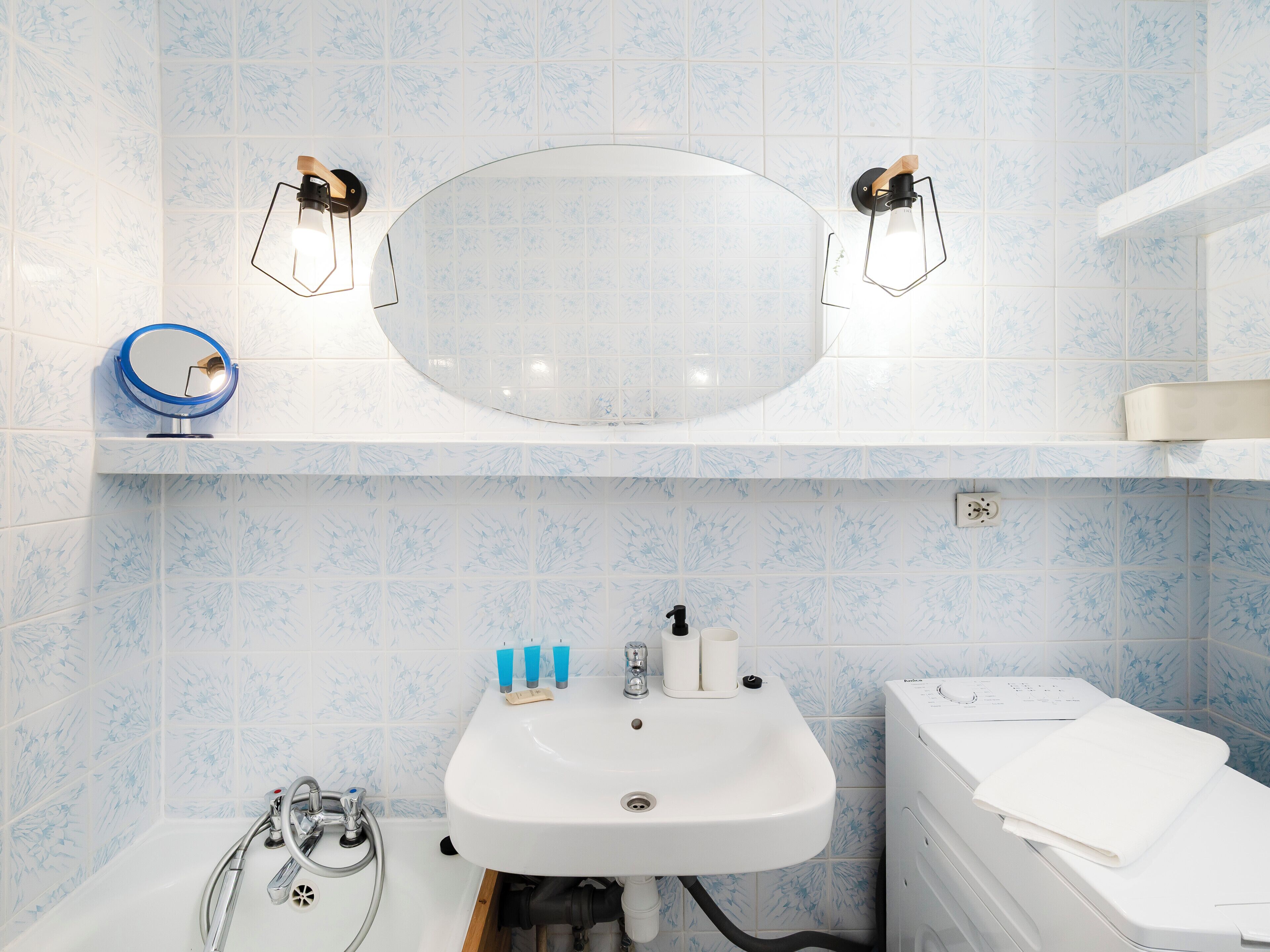 Ett badrum med ett vitt handfat, två väggmonterade lampor, en spegel och en vit tvättmaskin.
