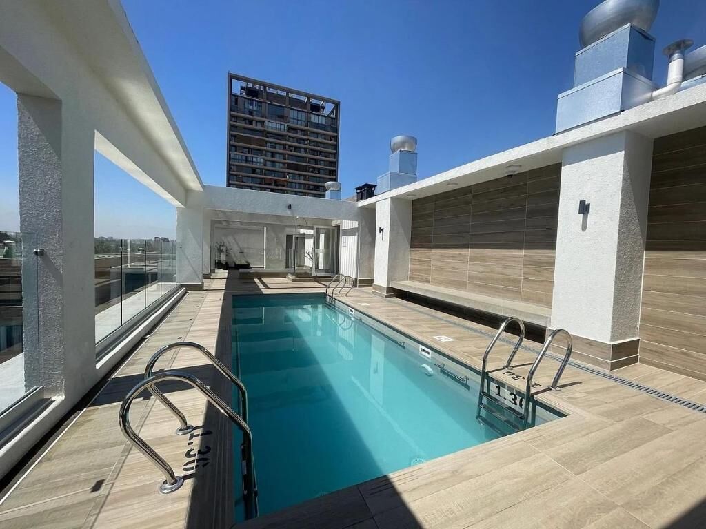Una piscina en la azotea con barandas de vidrio transparente, un diseño arquitectónico moderno y vistas a un rascacielos al fondo.