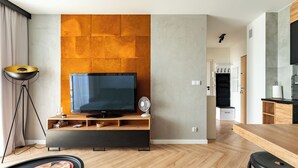 Una sala de estar moderna con una televisión de pantalla plana, piso de madera en espiga, una zona de comedor con mesa de madera, y una cocina al fondo.
