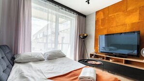 Una habitación de hotel moderna con una cama, un televisor, una lámpara de pie y vistas a edificios al exterior.