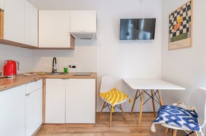 A compact kitchen with white cabinets, a red kettle, a sink, and a small table with two chairs. A television is mounted on the wall above the table. - Na Piasku Cozy Studio (Gliwice)