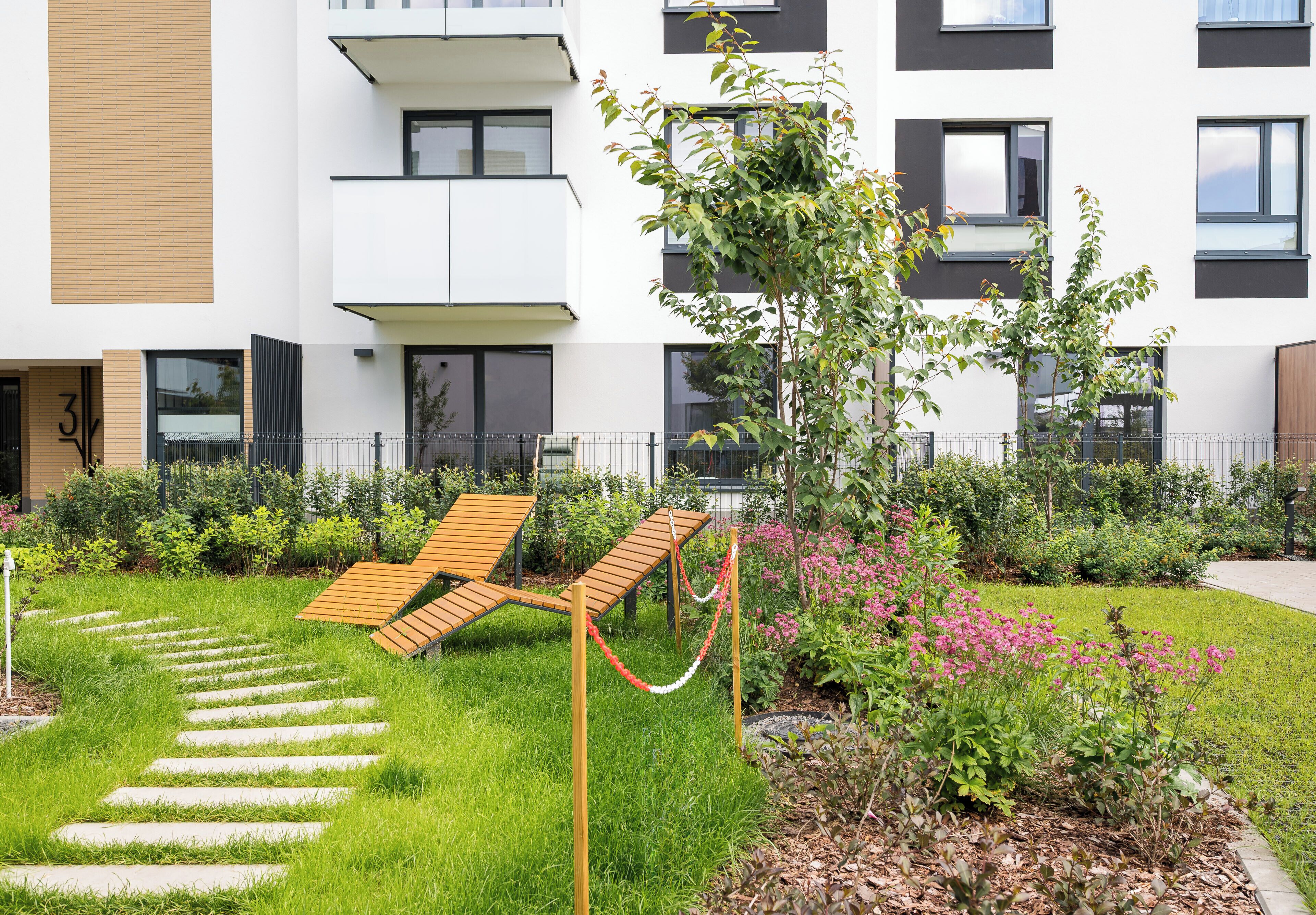 A modern building with a garden area featuring a wooden bench, a small tree, and pink flowers.