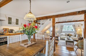 Een houten eettafel met een bloemenvaas, witte stoelen en een trap naar een woonruimte met een bank en een lamp.