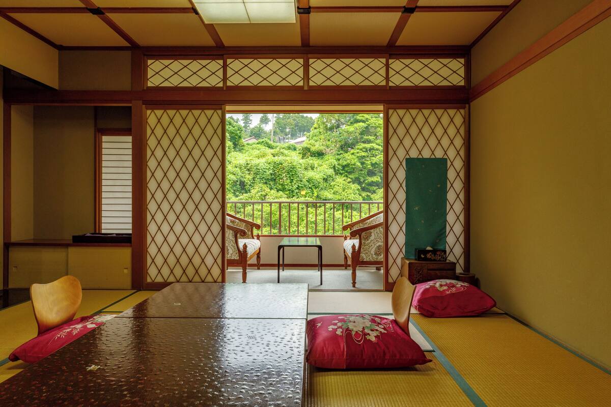 Um quarto japonês tradicional com tatames, uma mesa baixa e vista para um jardim exuberante através de uma porta deslizante.