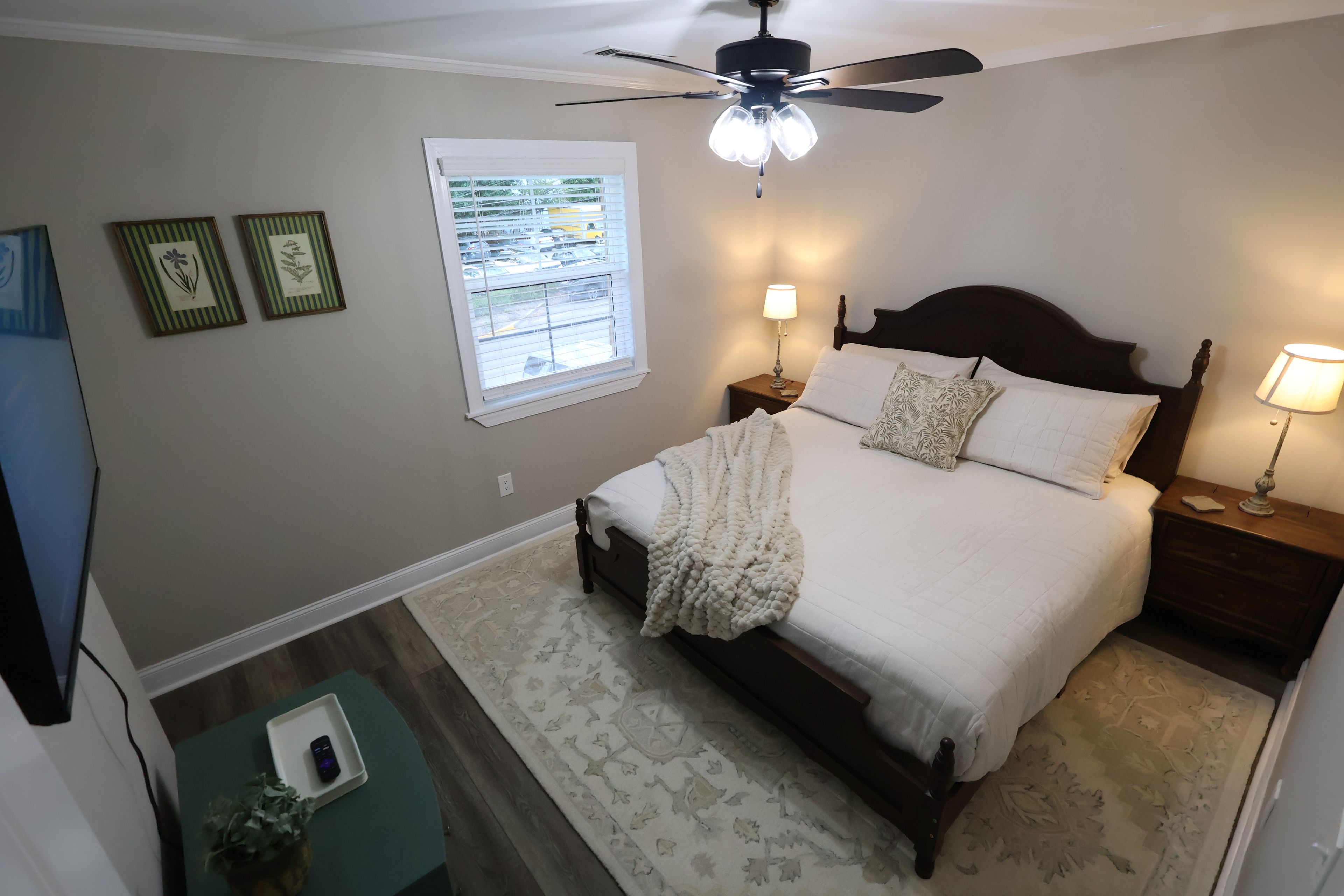 A bedroom with a bed, two bedside tables with lamps, a ceiling fan, a window with blinds, and a small table with a phone and a plant.