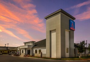 Um hotel moderno com um destaque para a placa 'Studio 6', estacionamento e céu claro.