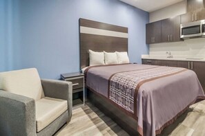 Quarto de hotel com cama, mesa de cabeceira, uma cadeira, cozinha compacta e parede azul.