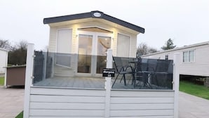 A mobile home with a covered deck, a sign indicating 'Morgana 10', and a patio area with chairs. - Ty Newydd Leisure Park 2-Bed Caravan, Sleeps 6 (Llanbedrgoch)