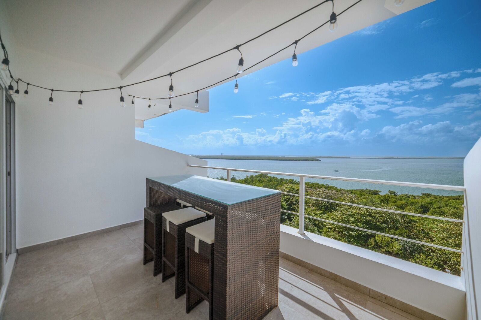 A balcony with a glass-top table and two chairs, overlooking a coastal view with a clear sky.
