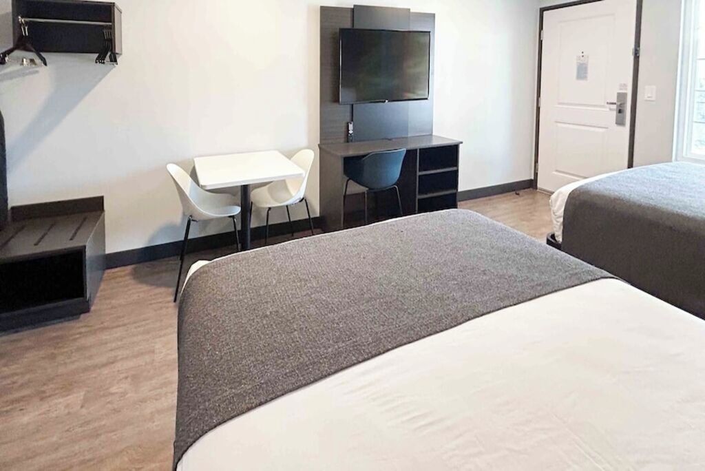 A hotel room with a bed, a desk with a chair, a TV mounted on the wall, and a door.