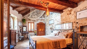 A rustic bedroom with a stone wall, wooden beams, a bed with a patterned bedspread, a desk with a chair, and a hanging light fixture. - Traditional apt in stone house in heart of Trogir (Trogir)