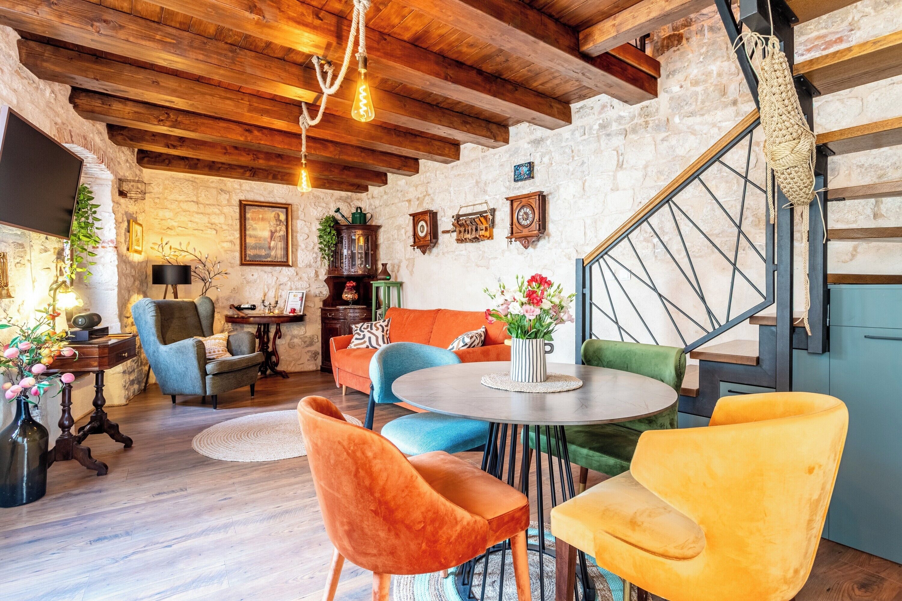 A rustic interior with exposed stone walls, wooden beams, a round table, and colorful chairs.