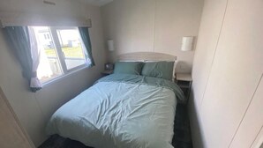 Ein kleiner, schmaler Raum mit einem Einzelbett, einem Fenster mit Vorhängen, einem Nachttisch und einer Wandleuchte.