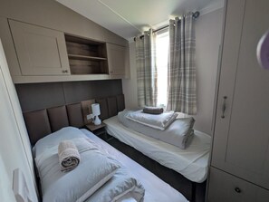Una habitación con dos camas, una mesita de noche con lámpara, un armario y una ventana con cortinas.
