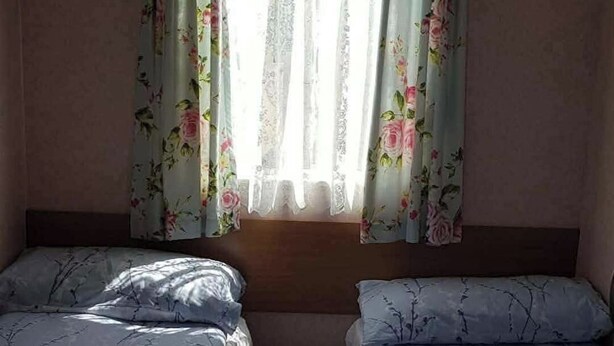 Huoneessa on kaksi sänkyä, ikkunassa on kukalliset verhot ja sängyn päädyssä on puinen koristepaneeli.