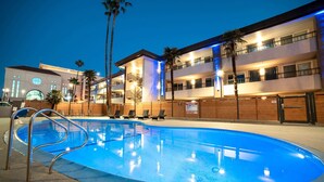 Une piscine avec des chaises longues, un immeuble doté de nombreux balcons et des palmiers.