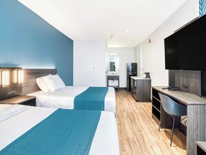 A hotel room with two beds, a desk, a chair, a TV, and a kitchenette.