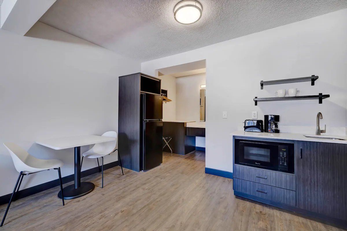Cocina pequeña y funcional con microondas, cafetera y un refrigerador compacto. Se observa una mesa de comedor con dos sillas y una pared blanca.