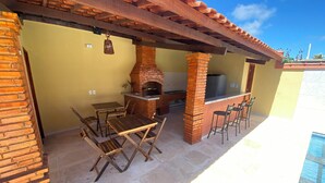 Um pátio externo com forno de tijolos, mesas e cadeiras de madeira, uma piscina e uma estrutura de pérgola.