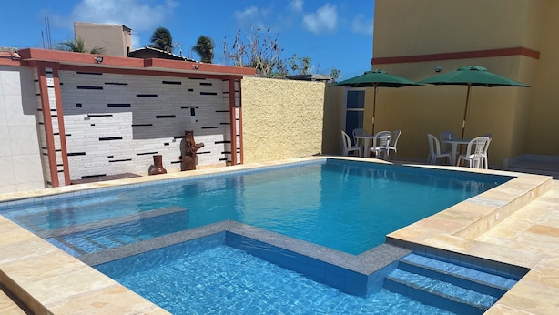 Piscina retangular com água azul cristalina, rodeada por azulejos bege e uma borda de pedra.