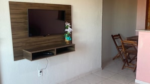 Uma sala de estar com uma TV de tela plana presa a um painel de madeira, uma mesa pequena com cadeiras e piso de azulejos.