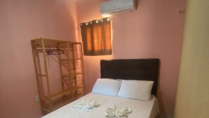 Uma cama individual com roupa de cama e toalhas brancas, um guarda-roupa de madeira, um candeeiro de teto e um aparelho de ar condicionado.