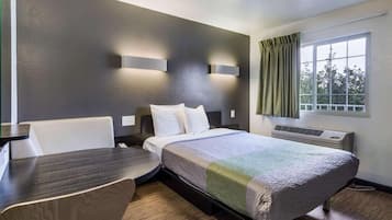 A hotel room with a bed, a desk, a chair, a window with curtains, and a wall-mounted air conditioning unit.