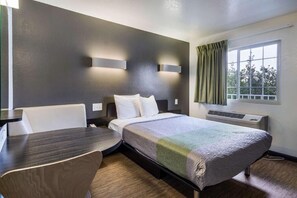A hotel room with a bed, a desk, a chair, a window with curtains, and a wall-mounted air conditioning unit.