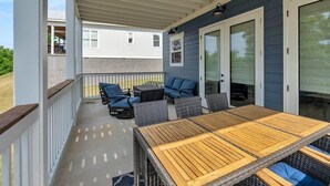 Een overdekt terras met een houten tafel en stoelen, een schuifdeur en uitzicht op een omliggend huis.
