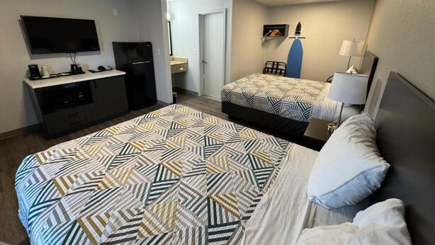Ein Hotelzimmer mit einem großen Bett, einem kleineren Bett, einem Schreibtisch mit Flachbildfernseher, einem Kühlschrank, einem Sessel und einer Lampe.