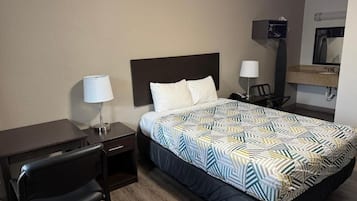 Habitación de hotel con cama, escritorio, silla, mesita de noche, lámpara, televisión y espejo.